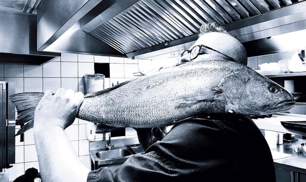 un homme tenant un gros poisson sur son épaule dans une cuisine de restaurant